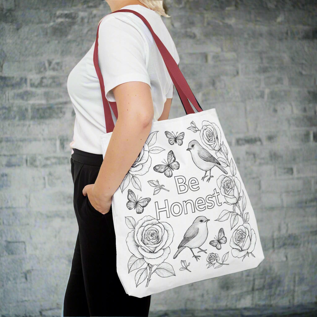 Person holding a white tote bag with floral and butterfly designs and the text 'Be Honest'.