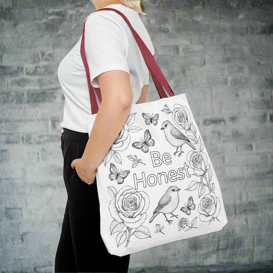 Person holding a white tote bag with floral and butterfly designs and the text 'Be Honest'.